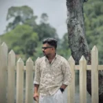 Man wearing a beige patterned shirt and sunglasses standing casually with one hand in pocket near a white picket fence and a large tree, with green foliage in the background.