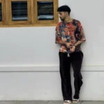 Man wearing a colorful tie-dye t-shirt, dark pants, and sandals standing against a white wall near a wooden window, looking to the side.