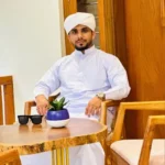 Man dressed in traditional white attire and turban sitting at a wooden table with sunglasses and a small potted plant.