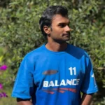 Man wearing a blue t-shirt with the word 'BALANCE' and the number 11 printed on it, standing outdoors with green foliage and flowers in the background.